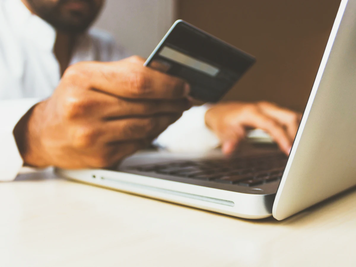 Persoană cu laptopul pe genunchi care ține în mână un card bancar, gata să plătească online
