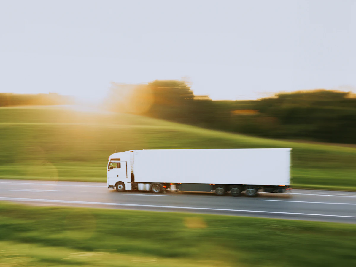 Camion alb cu semiremorcă pe un drum de țară, între câmpuri