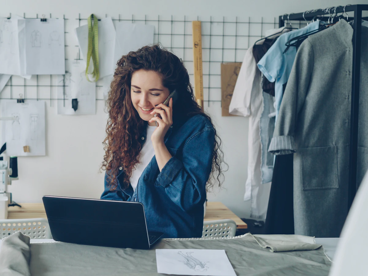 Designeriță de modă care vorbește la telefon și lucrează la laptop în atelierul ei